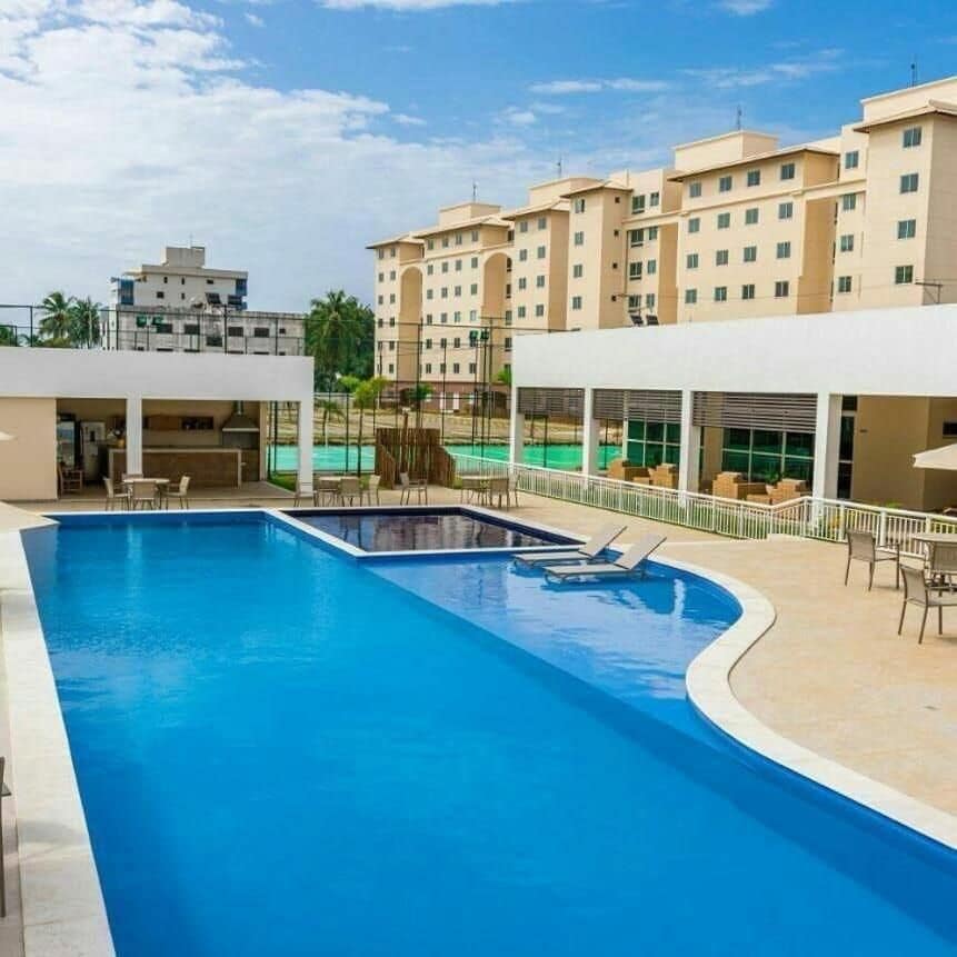A large swimming pool reflects the sky, surrounded by lounge chairs and shaded areas. In the background, tennis courts and other recreational facilities are visible, framed by modern buildings. Natural light enhances the serene setting for relaxation.