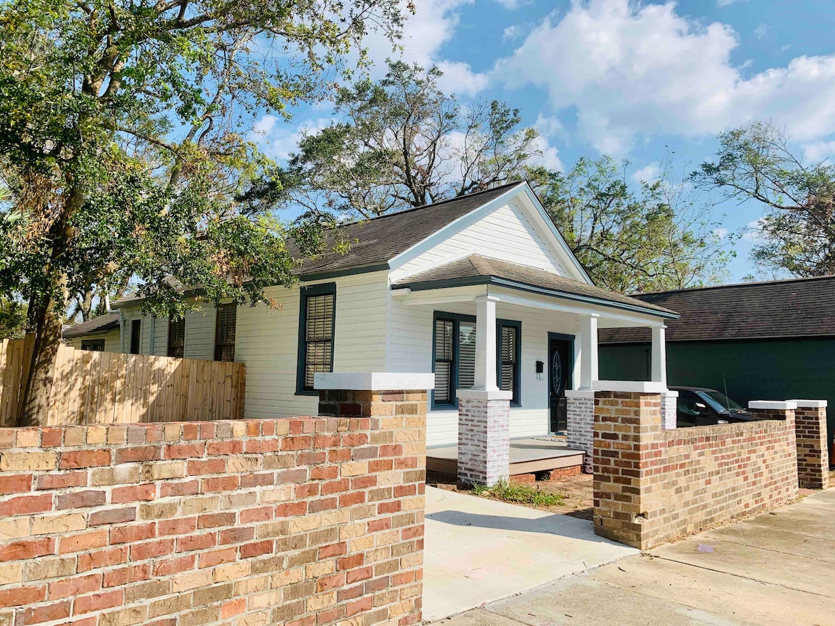 The exterior of a charming 1920's cottage is featured, showcasing a white façade with dark trim accents. A brick wall surrounds the front yard, and a spacious driveway is visible. Lush green trees provide shade in the background, and inviting steps lead to the front porch.