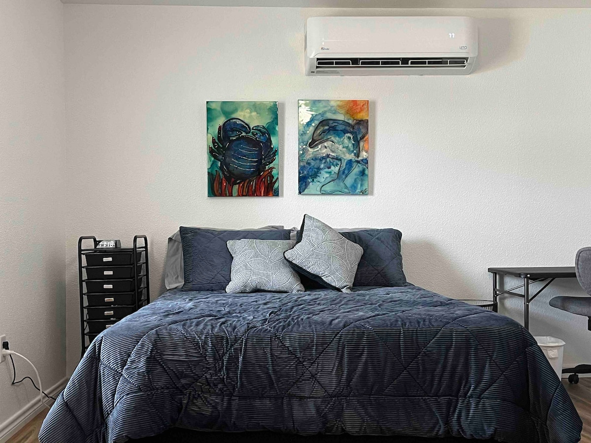 A spacious sleeping area is depicted, featuring a large bed dressed in dark bedding. Colorful wall art of sea creatures enhances the ambiance. An air conditioning unit is mounted above the bed, while a desk and a storage unit are visible in the room.