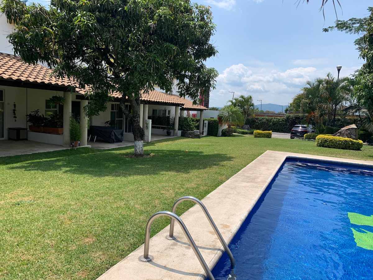 A spacious lawn is framed by neatly arranged gardens and a clear blue pool. The two-story house features a traditional tile roof, and visible are palm trees and manicured bushes. The inviting pool has a set of stainless steel steps leading into the water.