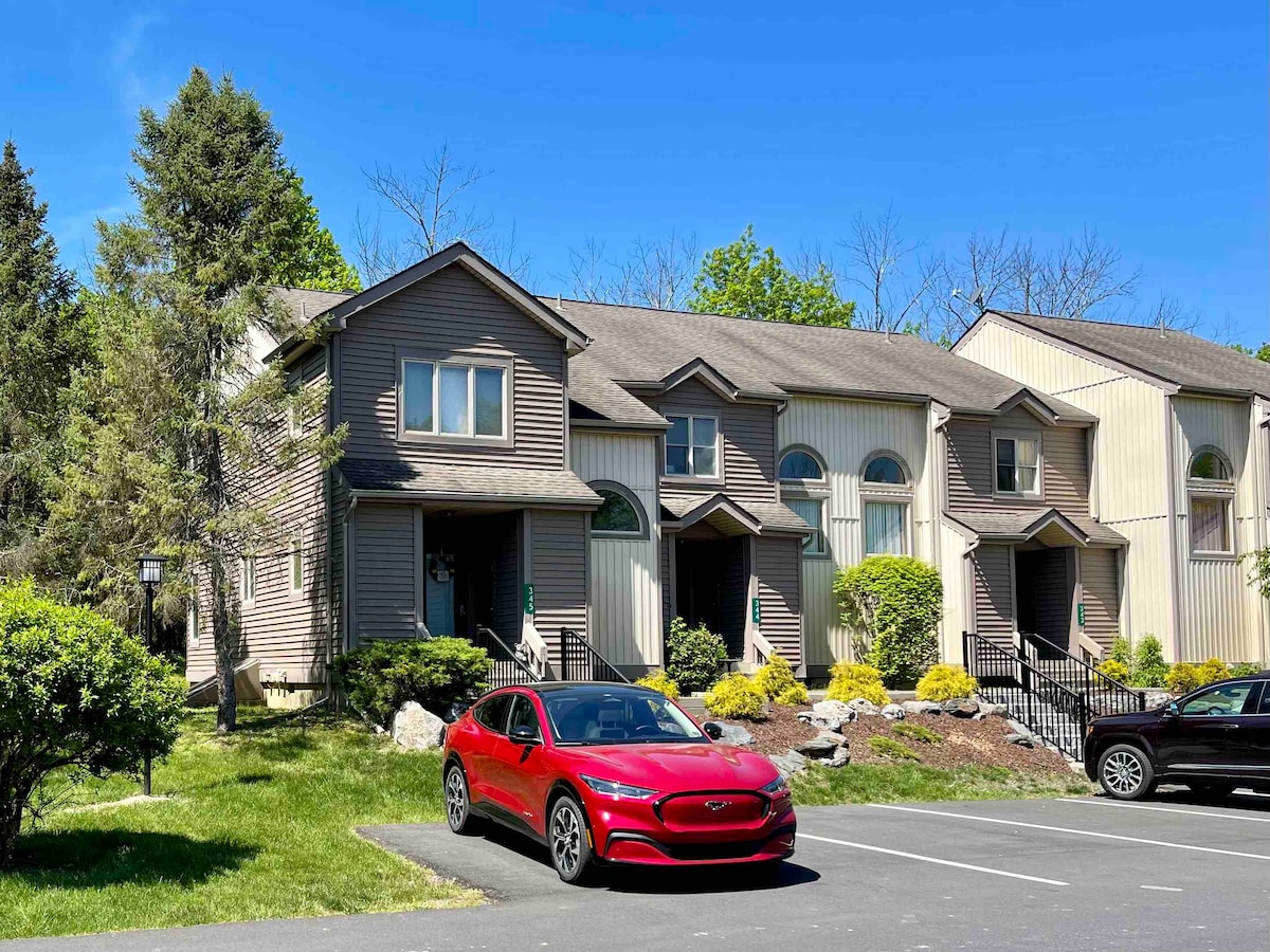 A modern townhouse is displayed against a clear blue sky, featuring large arched windows and well-maintained landscaping. A red car is parked in the designated spot directly in front of the house. The green lawn and garden beds add a touch of nature to the setting.
