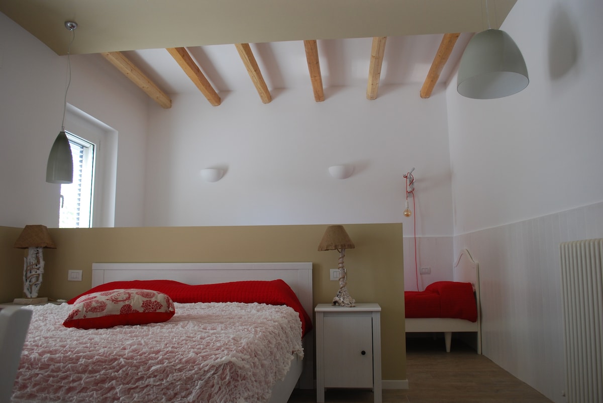 A spacious bedroom features a large bed with a textured red and white coverlet. Decorative lamps are positioned on either side of the bed, while a small twin bed is visible in the background. The ceiling showcases exposed wooden beams, and natural light enters through a window.