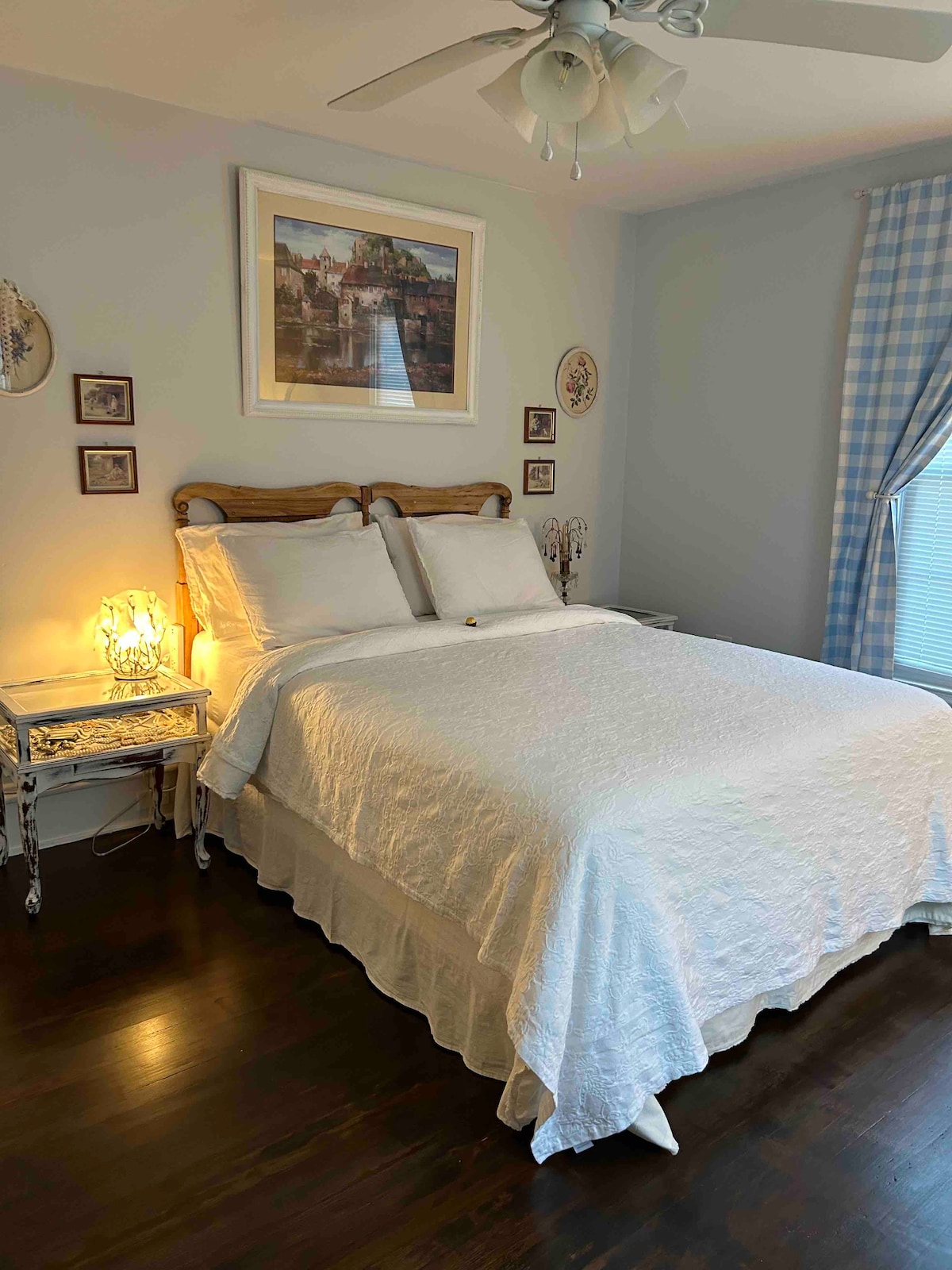 A queen bed with a white quilt is positioned against a light blue wall, complemented by two nightstands with decorative lamps. Framed artwork hangs above the bed, and light blue checkered curtains frame a window, creating a serene sleeping area.