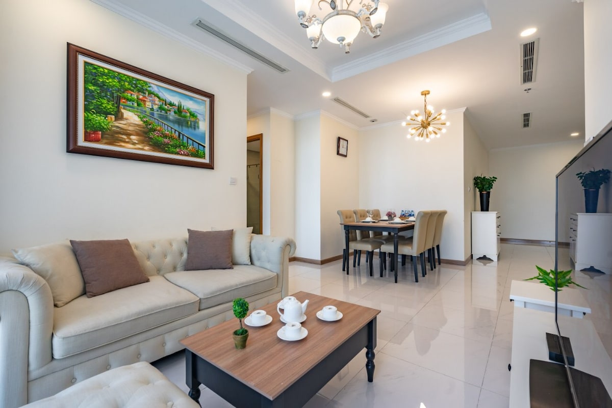 A modern living area features a light-colored sofa with decorative cushions, positioned beside a wooden coffee table set with tea ware. A large painting adorns the wall, and a dining area with beige chairs is visible in the background, illuminated by a stylish chandelier.