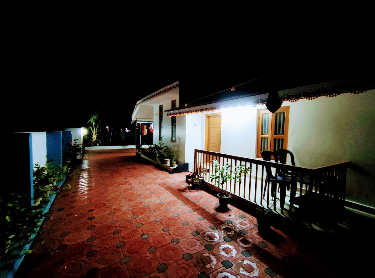 The outdoor area is illuminated softly by overhead lights, featuring a red-tiled path. A seating arrangement is visible on the porch, surrounded by potted plants. The structure displays traditional architectural elements, contributing to a serene nighttime ambiance.
