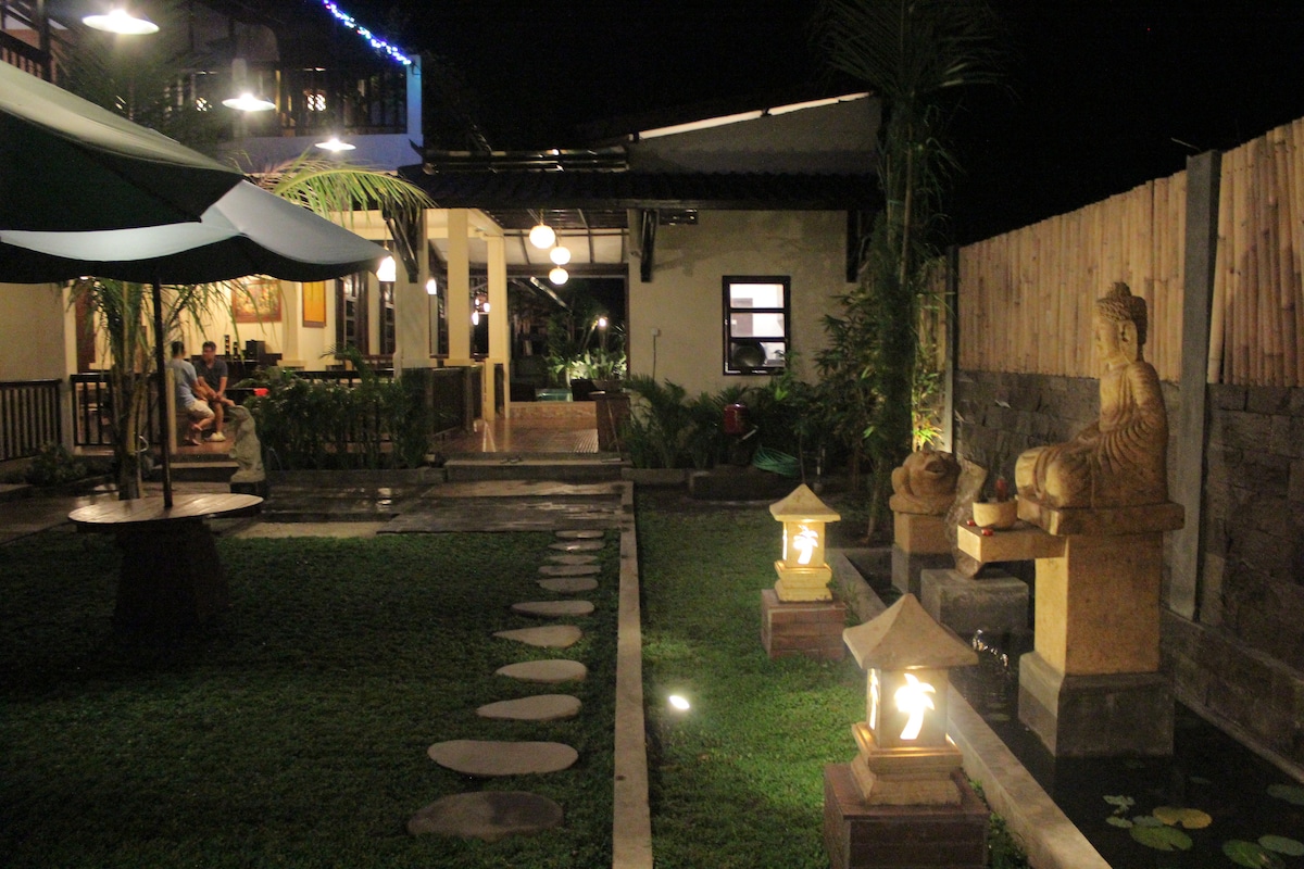 A serene outdoor area is highlighted by a manicured lawn, featuring stone paths that lead towards a tranquil statue of Buddha. Soft lighting illuminates decorative lanterns, while dining and lounge spaces are visible in the background, promoting a welcoming ambiance for guests.