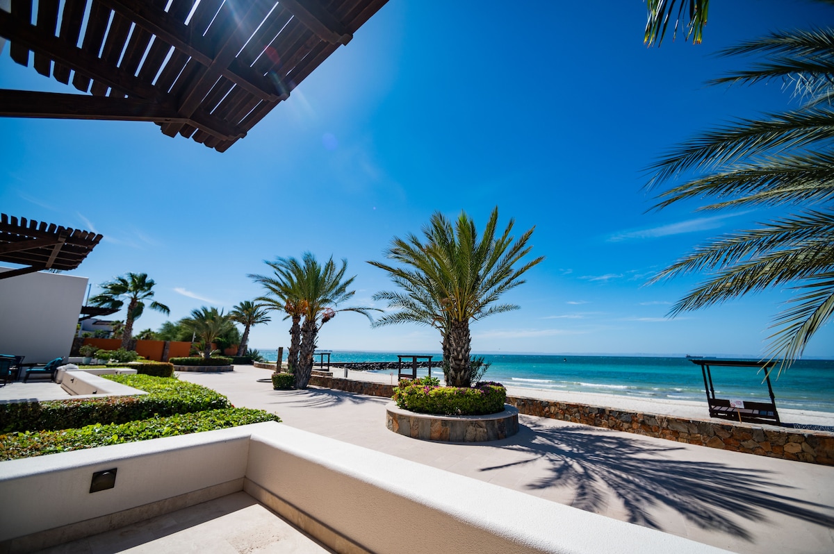 A spacious outdoor area is presented, featuring well-manicured gardens and palm trees lining the beachfront. The calm sea is visible in the background under a clear blue sky, inviting relaxation and tranquility. Comfortable seating areas enhance the coastal ambiance.
