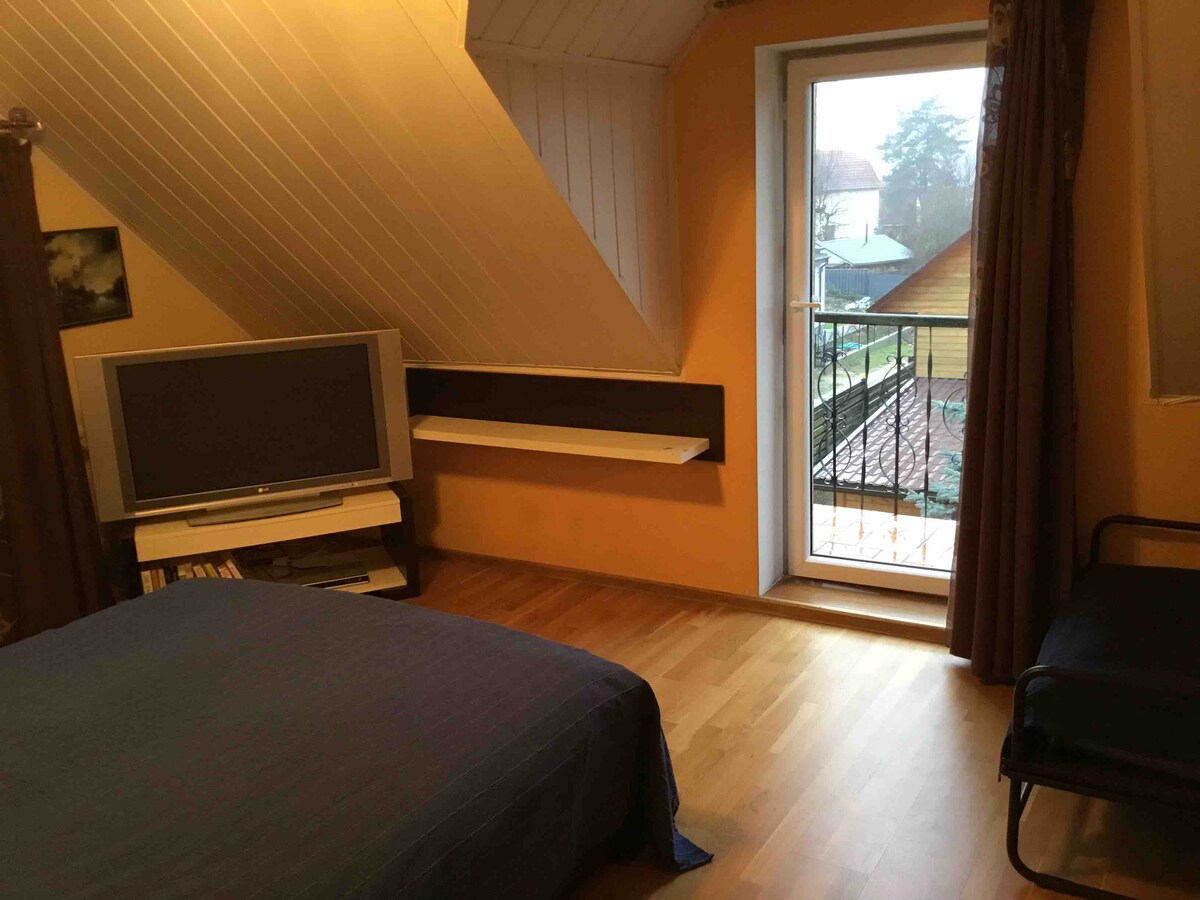 A room features angled wooden ceilings and warm orange walls. A spacious bed with blue bedding is positioned near a window that opens to a balcony. A television is mounted on a white shelf, and natural light fills the space from the door leading outside.