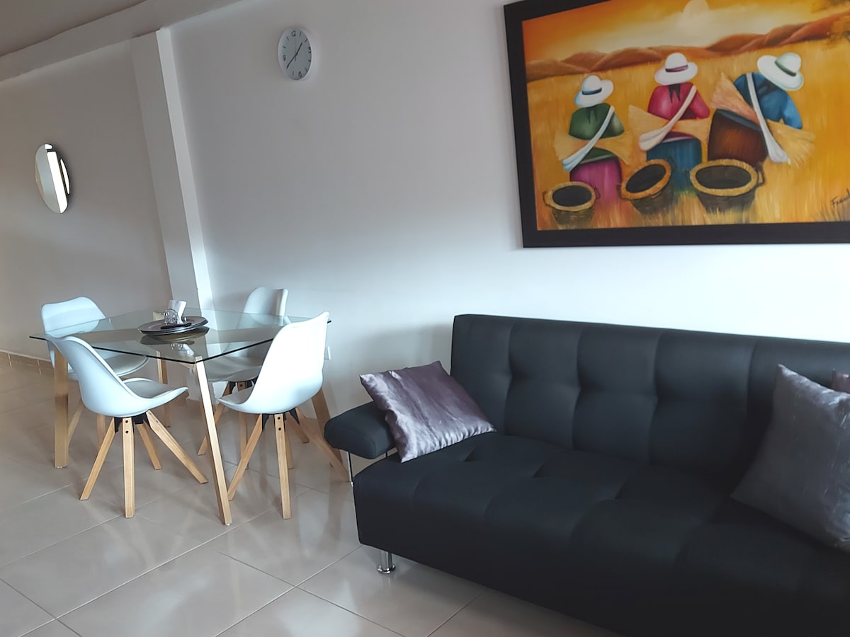 A versatile living space features a dark sofa and a glass dining table surrounded by white chairs. A colorful wall painting depicting three figures is prominently displayed. Natural light brightens the area, while a wall clock adds a functional touch.