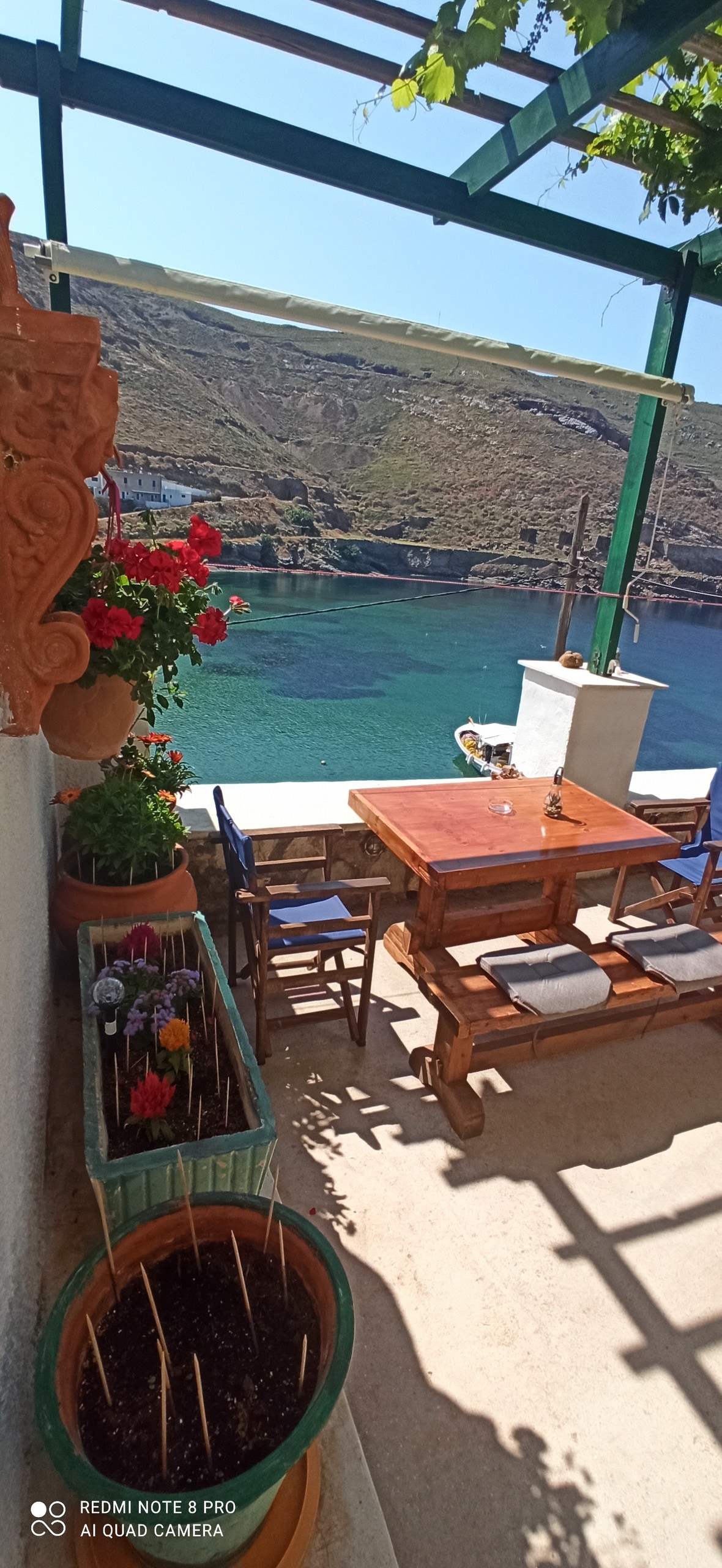 A dining area features a wooden table surrounded by chairs on a shaded terrace. Potted flowers add color, while views of the clear sea and surrounding hills create a serene atmosphere. A trellis overhead provides partial shade, enhancing the outdoor space.