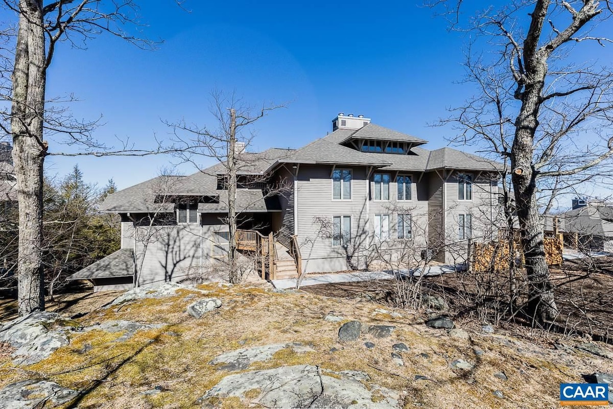 The exterior of a multi-level building is showcased against a clear blue sky. Natural stone surfaces and wooden decks are visible, surrounded by leafless trees. A walking path leads to the entrance, enhancing accessibility to this mountain retreat.