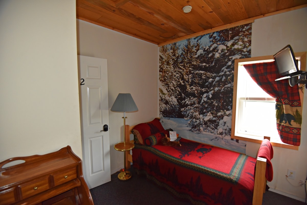 A cozy room features a single XL bed adorned with a patterned red blanket. A scenic mural of a snowy landscape is displayed on one wall, while a small desk accompanies a lamp and a window dressed with a plaid curtain. A wooden dresser is also present.