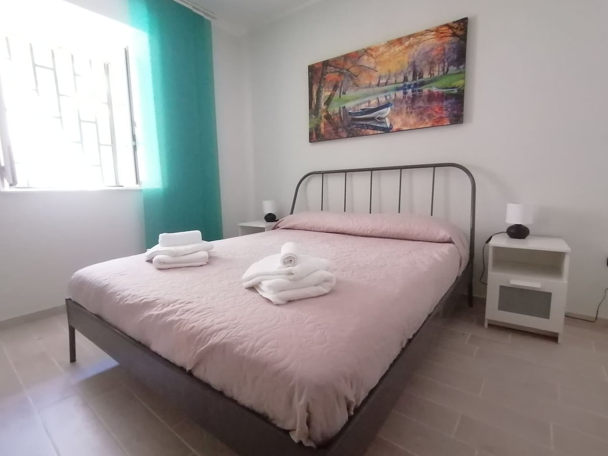 A cozy bedroom features a spacious bed with a light pink quilt and neatly arranged white towels. A colorful painting is displayed above the bed, while natural light filters through a window on the side, enhancing the serene atmosphere.