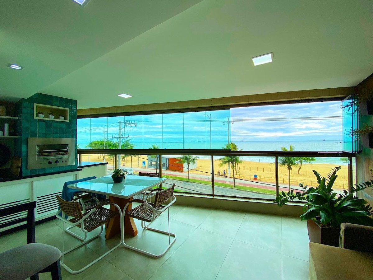 A modern dining area features a glass wall offering unobstructed views of the beach. A circular table, surrounded by four chairs, is complemented by a potted plant. Natural light fills the space, enhancing the serene ambiance with the ocean visible outside.
