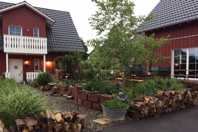 A charming exterior view of a red, two-story building framed by lush greenery and decorative stonework. The entrance is adorned with a small balcony, and various seating areas are visible among garden features. Wood piles are neatly stacked along the pathway.