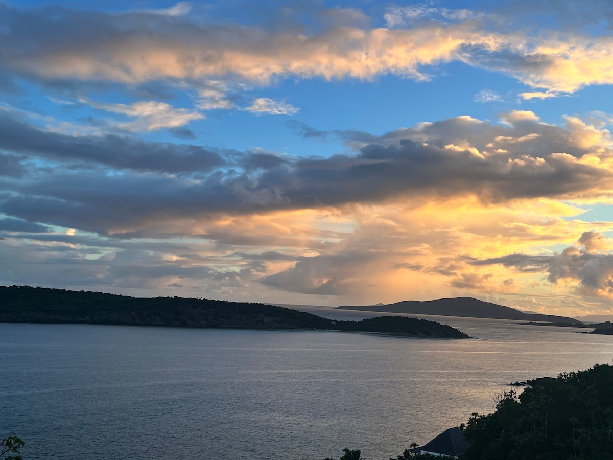 The image captures a panoramic view of the Atlantic Ocean, framed by rolling hills and distant islands. Soft clouds mingle with hues of orange and blue during sunset, reflecting off the water's surface. The scene evokes a peaceful coastal atmosphere.