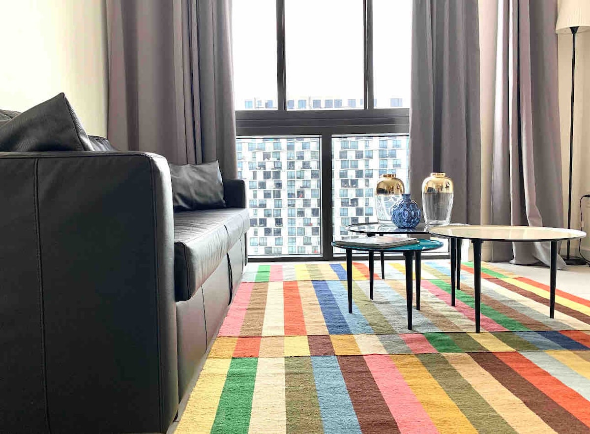 A modern living area features a black sofa and a striped area rug in various colors. Two coffee tables, one round and one oval, are positioned in front of the sofa. Floor-to-ceiling windows provide natural light, framed by long curtains.