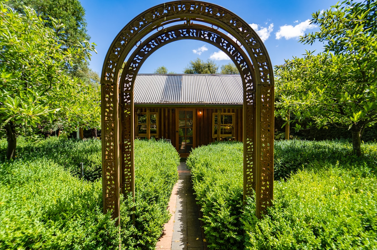 A quiet pathway is bordered by lush greenery, leading to the entrance of Glebe Cottage. The charming structure features wooden exterior walls and a metal roof, framed by vibrant trees under a clear blue sky.