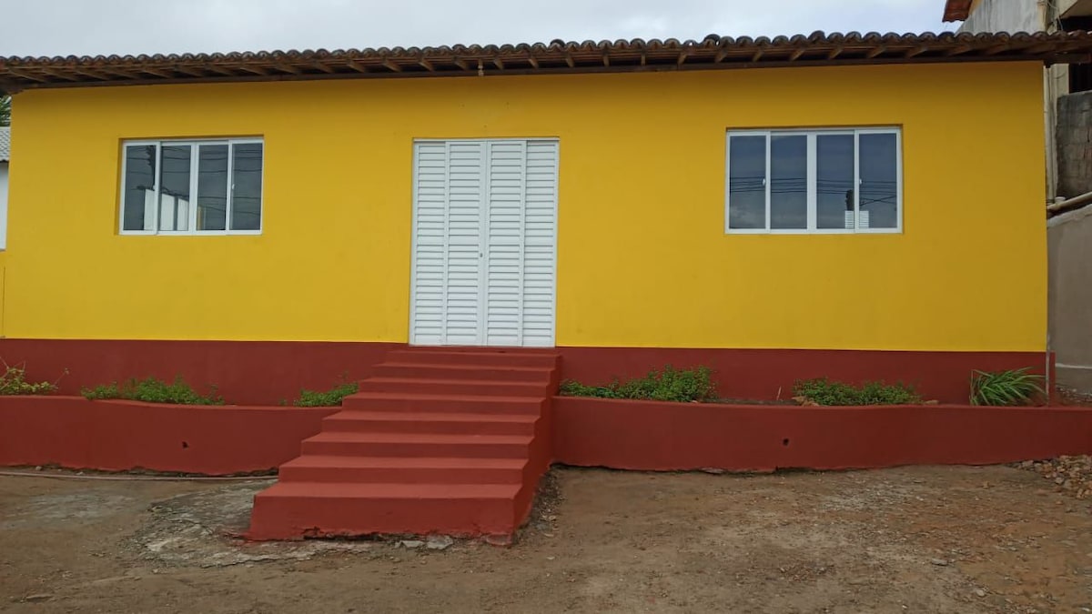 A vibrant yellow exterior of the house is featured, highlighted by three large windows offering natural light. A set of red steps leads to a white double door entrance, surrounded by a low planter with greenery. The roof is supported by wooden beams.