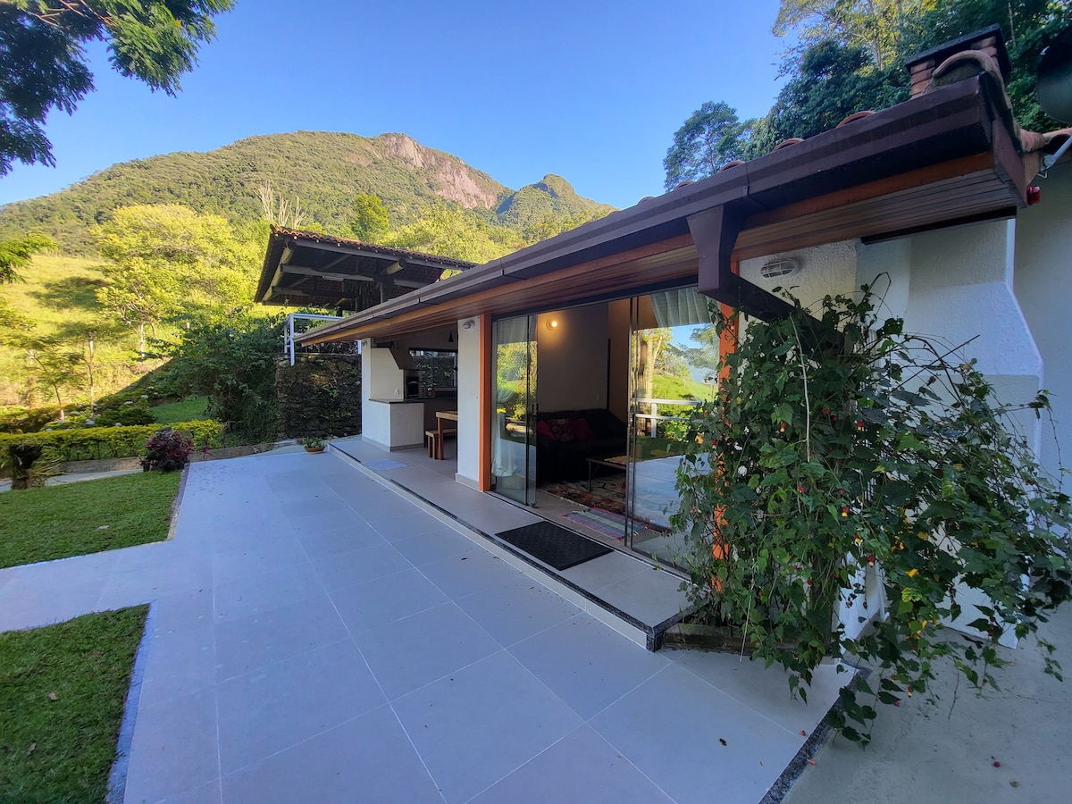 The exterior of the chalet features large glass sliding doors that open to a spacious patio. Lush greenery surrounds the structure, with mountains visible in the background, providing a natural and serene landscape. The pathway is lined with carefully maintained grass.