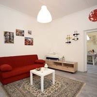 A bright living area showcases a red sofa complemented by a light-colored coffee table, positioned atop an intricately patterned rug. Walls are adorned with various framed photographs, adding visual interest. A doorway in the background leads to a visible kitchen space.