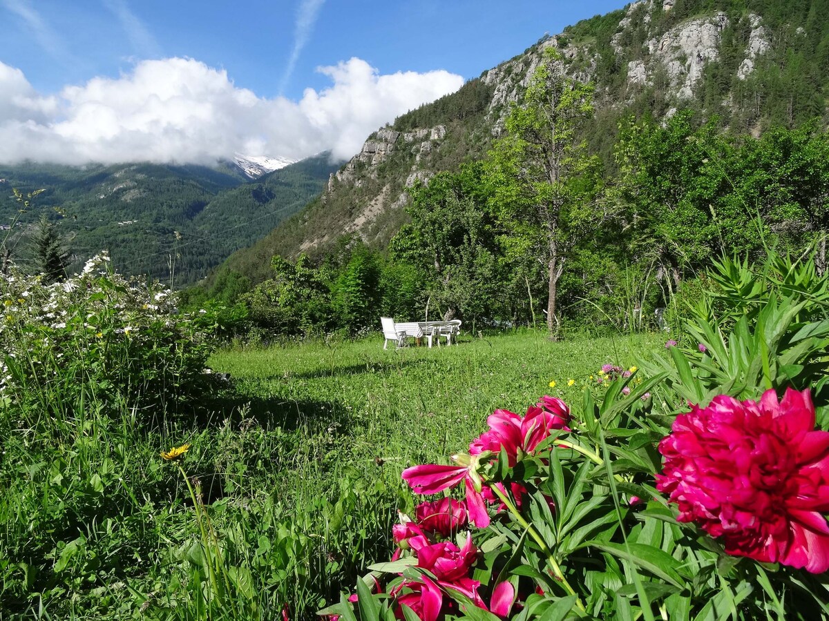 A serene garden scene is presented, featuring vibrant flowers in the foreground and a white seating set in the background. The lush green landscape stretches to meet the mountains under a partly cloudy sky, creating a peaceful outdoor atmosphere.
