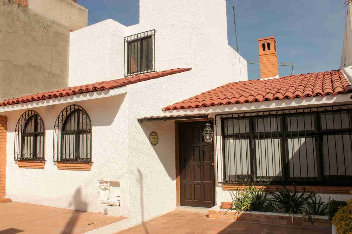 A single-story house features a white exterior with a red-tiled roof and decorative arches over the windows. A wooden front door is complemented by a lantern on the wall. Lush greenery is visible in the front yard, adding a fresh touch.