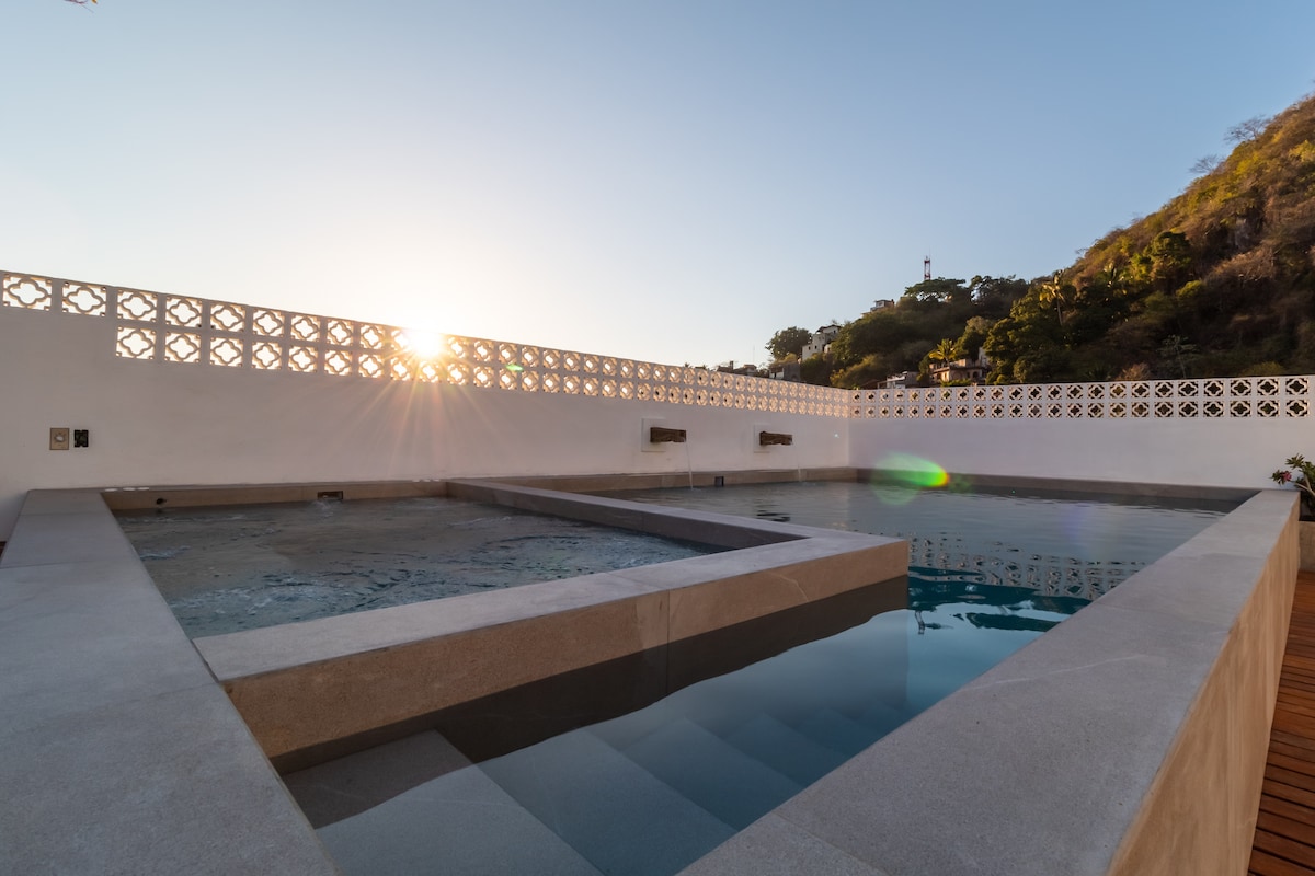 A shared rooftop area features a refreshing pool with steps leading down into the water. The gentle sun begins to set, casting a warm light over the space. The surrounding wall includes decorative patterns, contributing to the serene atmosphere.