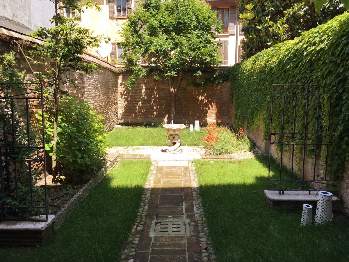 A private garden area features well-maintained grass and a central stone fountain. Borders of climbing plants and shrubs create a secluded ambiance. Brick walls provide structure and a sense of privacy, while sunlight filters gently through the greenery, illuminating the serene space.
