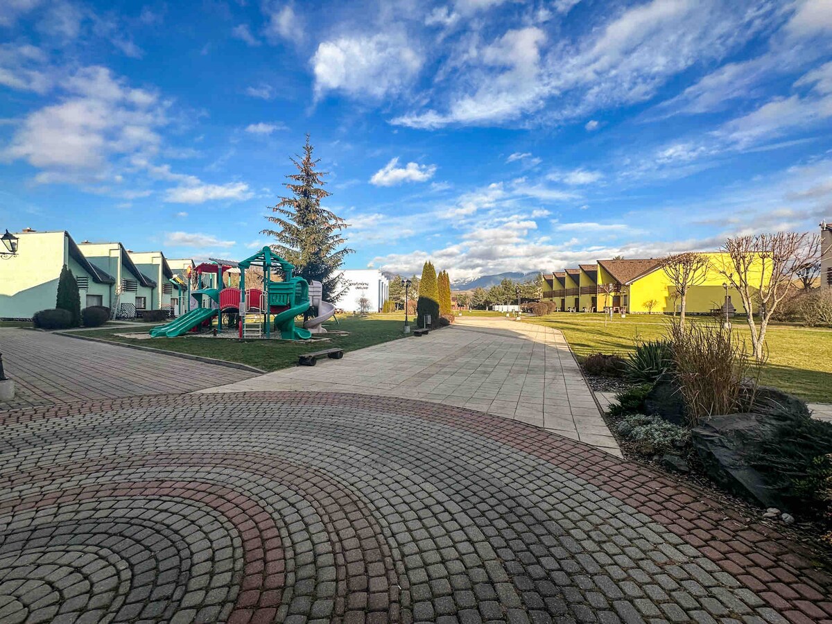 A spacious outdoor area is visible, featuring a playground with a slide and climbing structure. Greenery and neatly arranged flower beds provide a natural touch, while various colored buildings form a backdrop under a clear blue sky with scattered clouds.