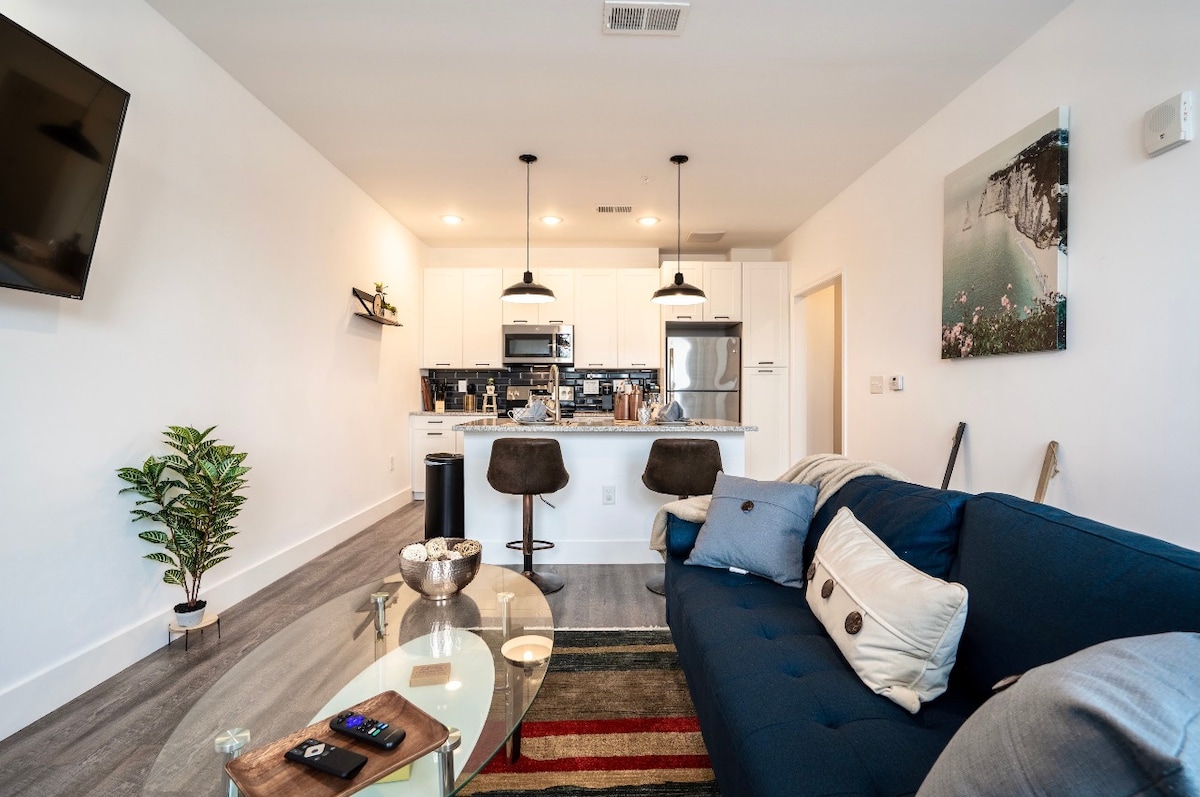 A modern living area features a blue sofa, an elegant glass coffee table, and a small potted plant. The kitchen showcases white cabinetry, high-end appliances, and a central island with bar seating, all under warm pendant lighting. A flat-screen TV is mounted on the wall.