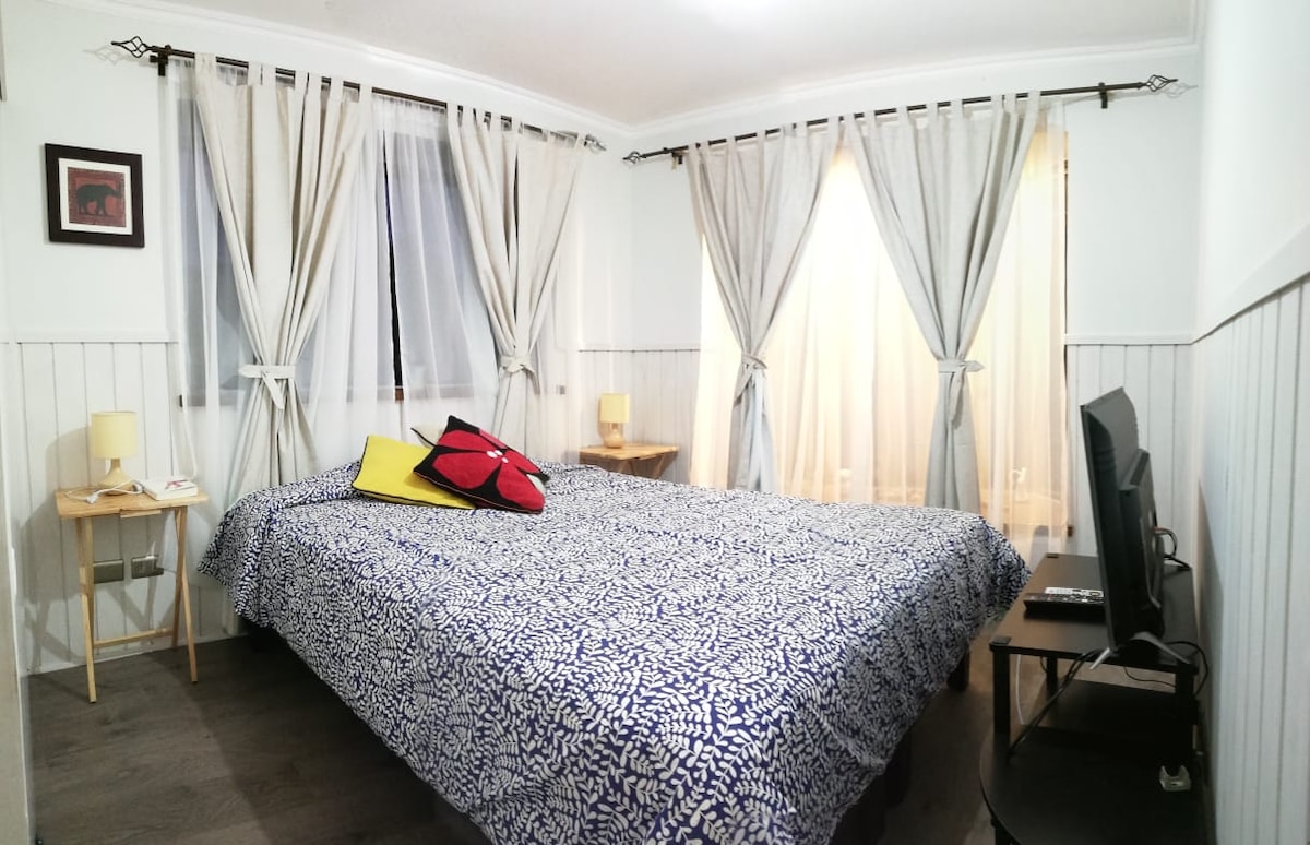 The bedroom features a spacious bed covered with a patterned blue and white blanket. Two bedside tables are situated beside the bed, each holding a lamp. Natural light filters through sheer curtains, complementing the soft color palette of the room.