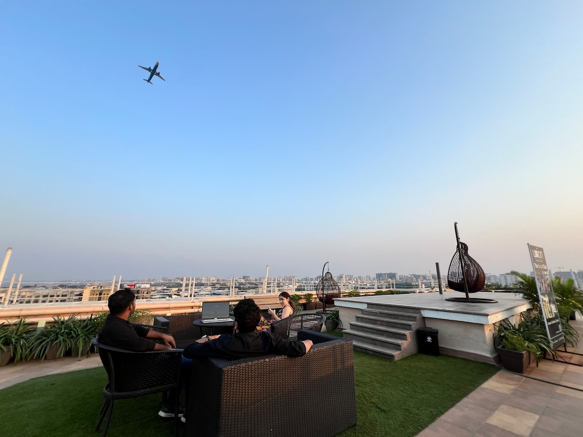A rooftop terrace offers a spacious setting with comfortable seating arranged around a low table. Lush green plants line the perimeter, while an airplane is seen flying overhead against a clear sky. Steps lead up from the terrace to an elevated area.