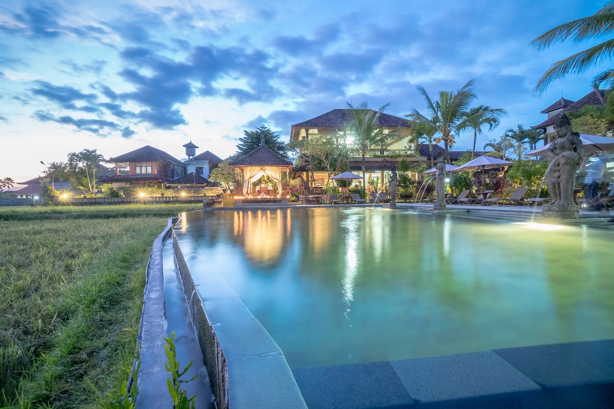 A serene swimming pool is set against a twilight sky, reflecting the soft glow of surrounding lights. Lush greenery and palm trees border the pool area, with comfortable lounge seating and traditional structures visible in the background.