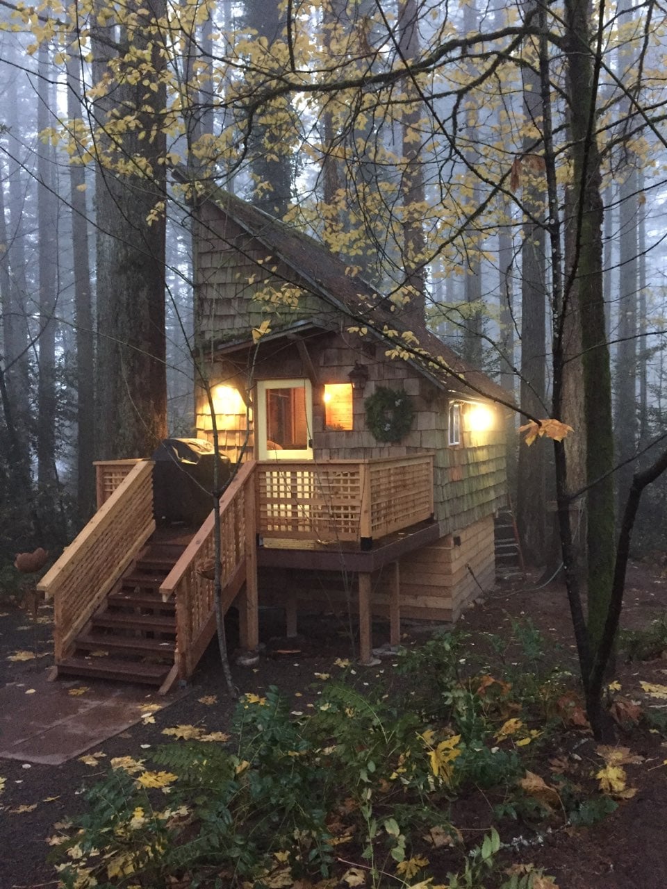 The cabin's exterior features a rustic design with wooden shingles, complemented by a welcoming wreath on the door. Soft lighting illuminates the entrance, while a staircase leads to the deck surrounded by tall trees and fallen leaves, creating a serene forest atmosphere.