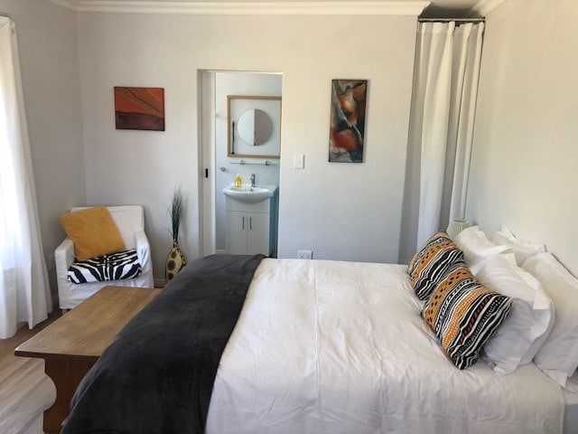 A bright and airy bedroom features a spacious bed adorned with patterned pillows. A small table is positioned beside the bed, and a comfortable chair is placed in the corner. Art pieces are displayed on the walls, and a bathroom is visible through an open doorway.