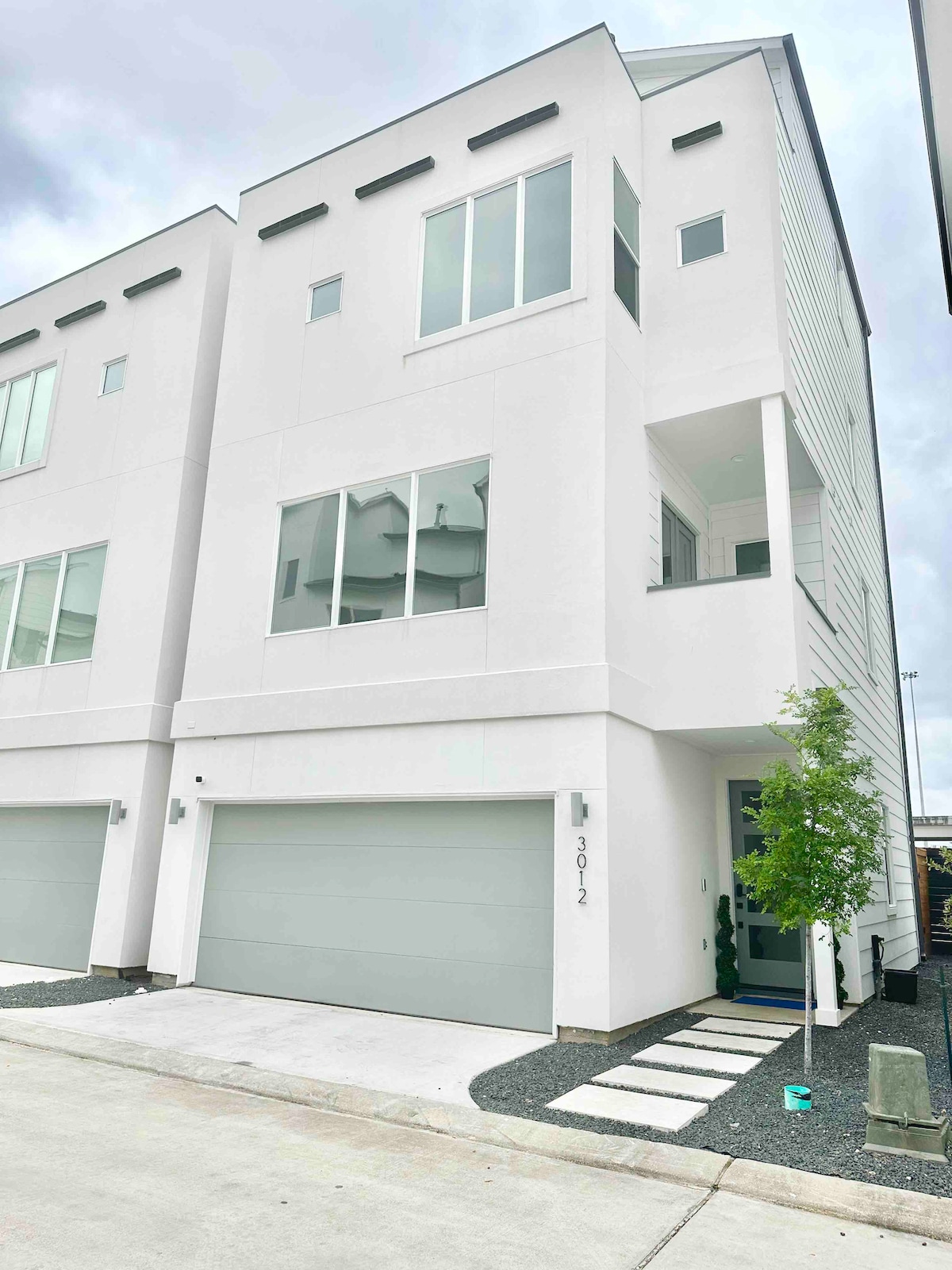 The exterior showcases a modern three-story home with a smooth white façade and large windows. A clean driveway leads to a spacious garage. A small tree and a stone pathway add a touch of greenery, enhancing the welcoming appearance of the property.