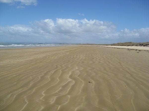 A wide expanse of golden sand stretches along the beach, with gentle undulations marking its surface. The ocean waves can be seen in the distance, and a clear blue sky with scattered clouds creates a serene ambiance.