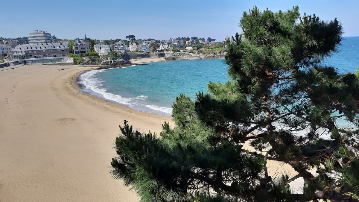 A sandy beach curves along a crescent coastline, framed by sparse greenery. Calm turquoise waters gently lap against the shore, with a small residential area visible in the background, nestled among trees. The scene captures a serene atmosphere, reflecting a stunning natural environment.