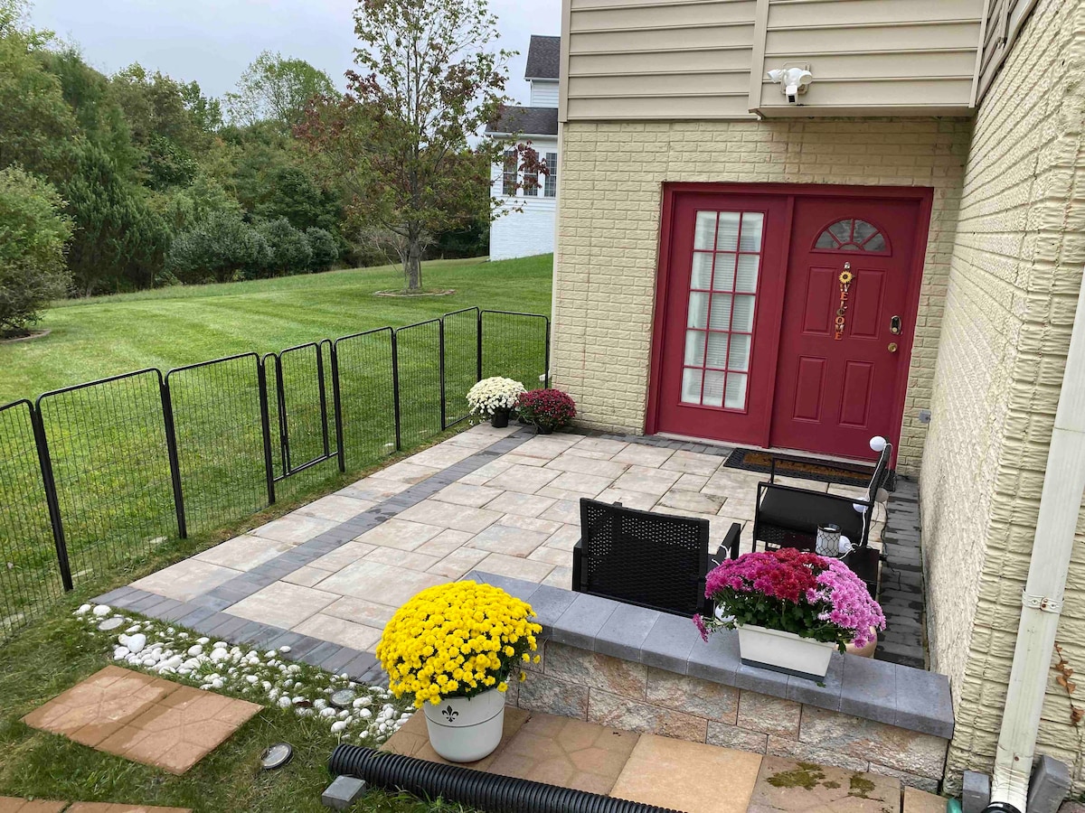 A private patio area features a spacious layout, with a red door leading into the suite. Potted flowers, including vibrant chrysanthemums, add color to the scene. A sectioned fence surrounds the patio, creating a cozy outdoor sitting area on patterned stone flooring.