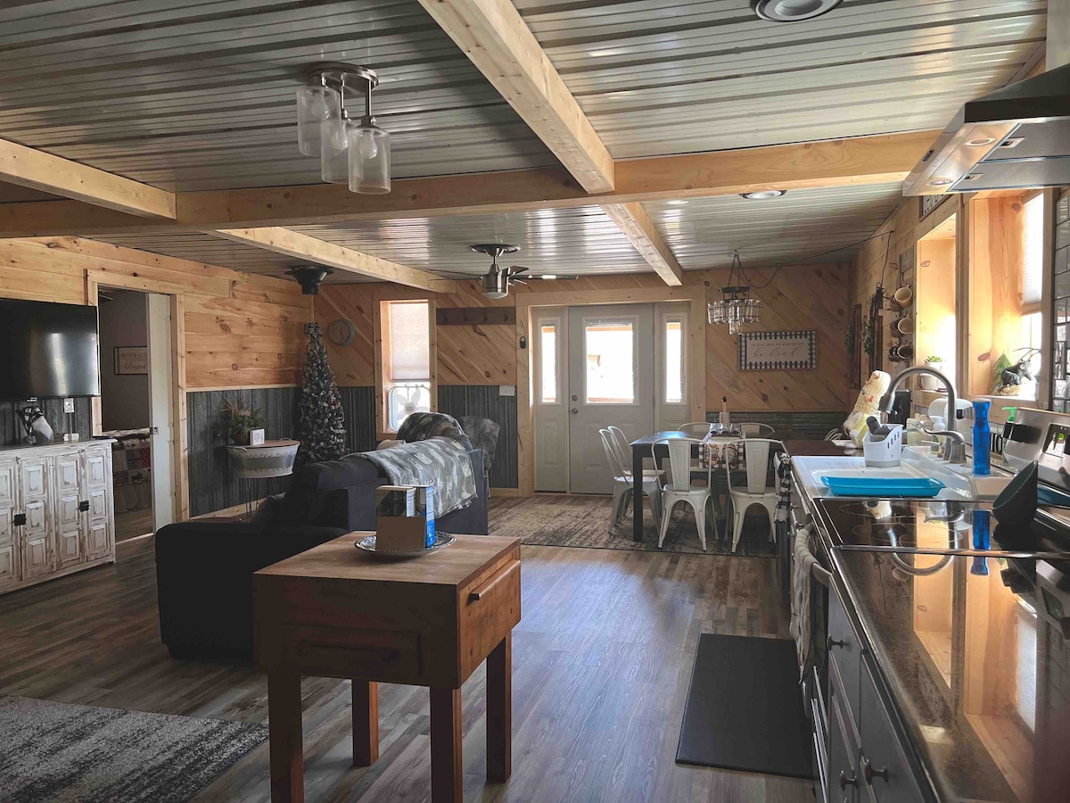 An open-plan living space features wooden ceiling beams and a combination of light wood and gray paneling. A cozy seating area with a dark couch is complemented by a dining table set for six. Natural light floods in through multiple windows.