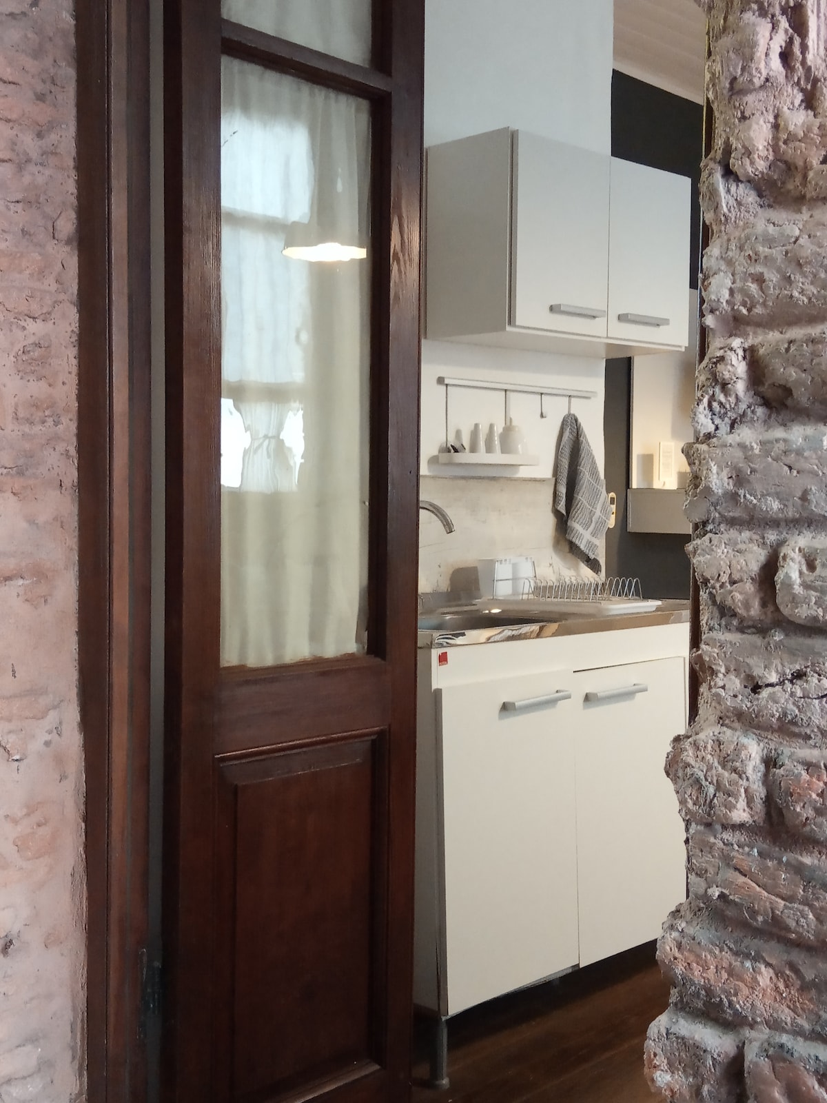 A small kitchenette is visible through a wooden door. The area includes white cabinets, a sink, and basic kitchenware displayed on a countertop. Exposed brick walls are present, adding texture to the space. Natural light enters softly through a nearby window.