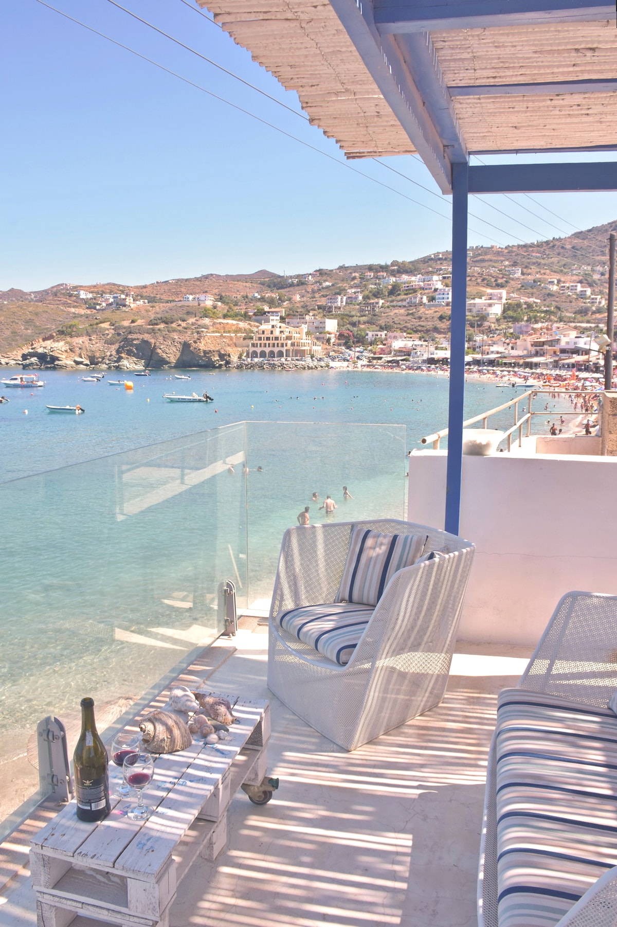 A private terrace overlooking a calm bay features comfortable seating along with a small table. The clear water reflects the surrounding hills and nearby beach activity, with boats gently bobbing on the surface. A picturesque coastal village is visible in the background.