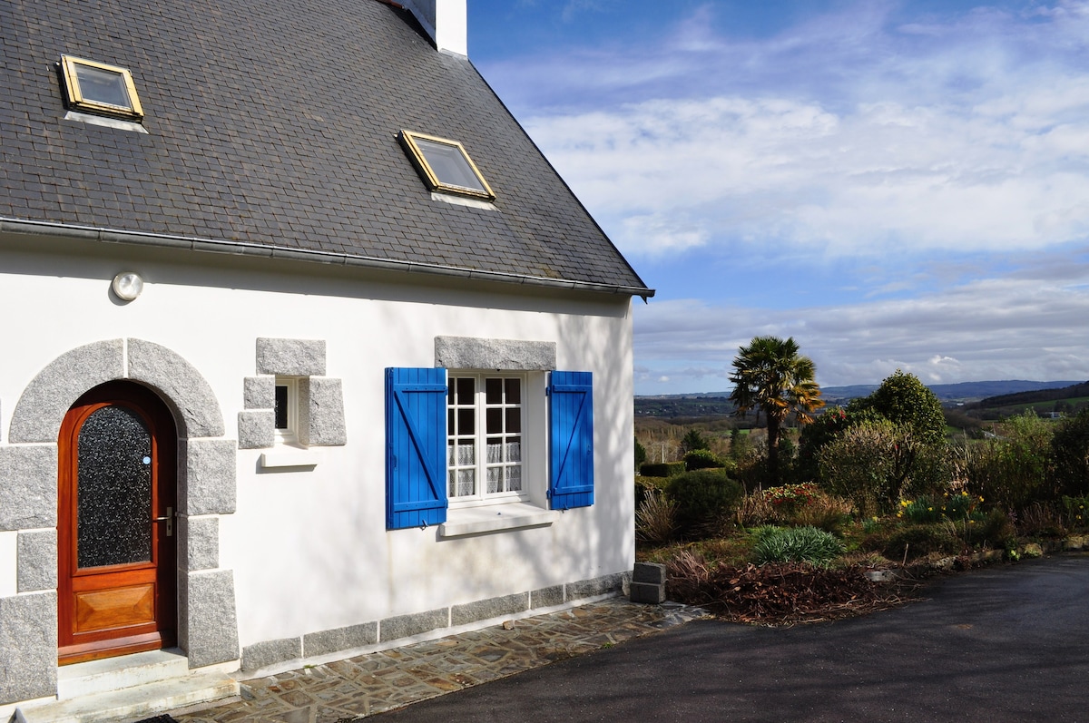 The exterior of a charming house is presented, featuring a gray slate roof and white walls accentuated by blue shutters. A wooden door framed with stone leads to the interior, while landscaped gardens and distant hills are visible in the background.