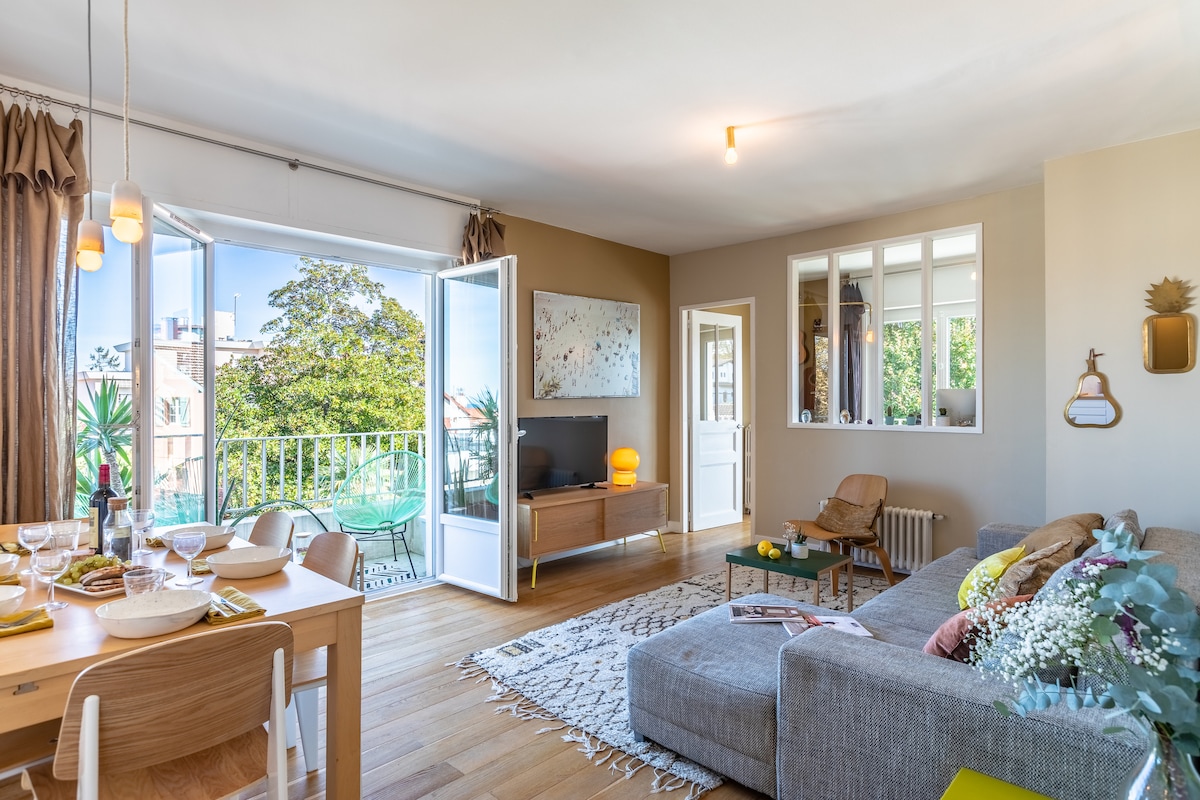 A bright living space is shown with large windows opening to a balcony. Natural light fills the room, highlighting a comfortable gray sectional sofa and a dining table set for a meal. Decorative elements include a rug and plants, enhancing the inviting setting.