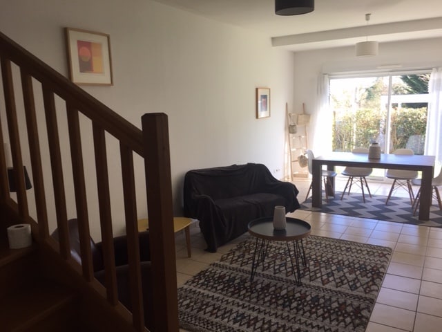 A bright living area features large windows overlooking an enclosed garden. A wooden dining table is surrounded by white chairs. A dark sofa and armchair are positioned on a patterned rug, while a small table with a decorative item adds a touch of detail.