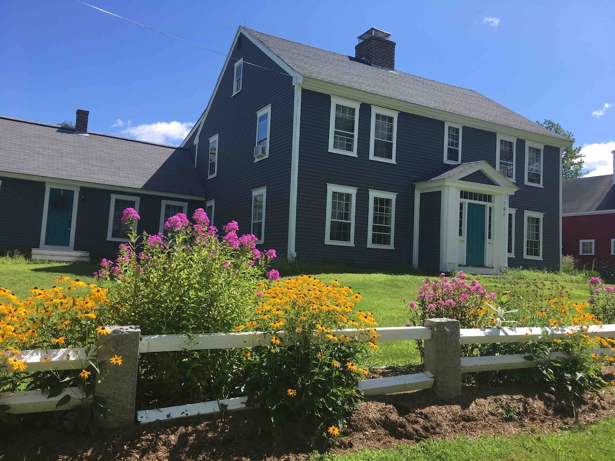 The exterior of a traditional New England-style farm house is displayed, featuring a dark blue façade and white trim. A manicured lawn with vibrant yellow and pink flowers surrounds a white fence, adding a colorful touch to the rural landscape. A clear blue sky enhances the scene.