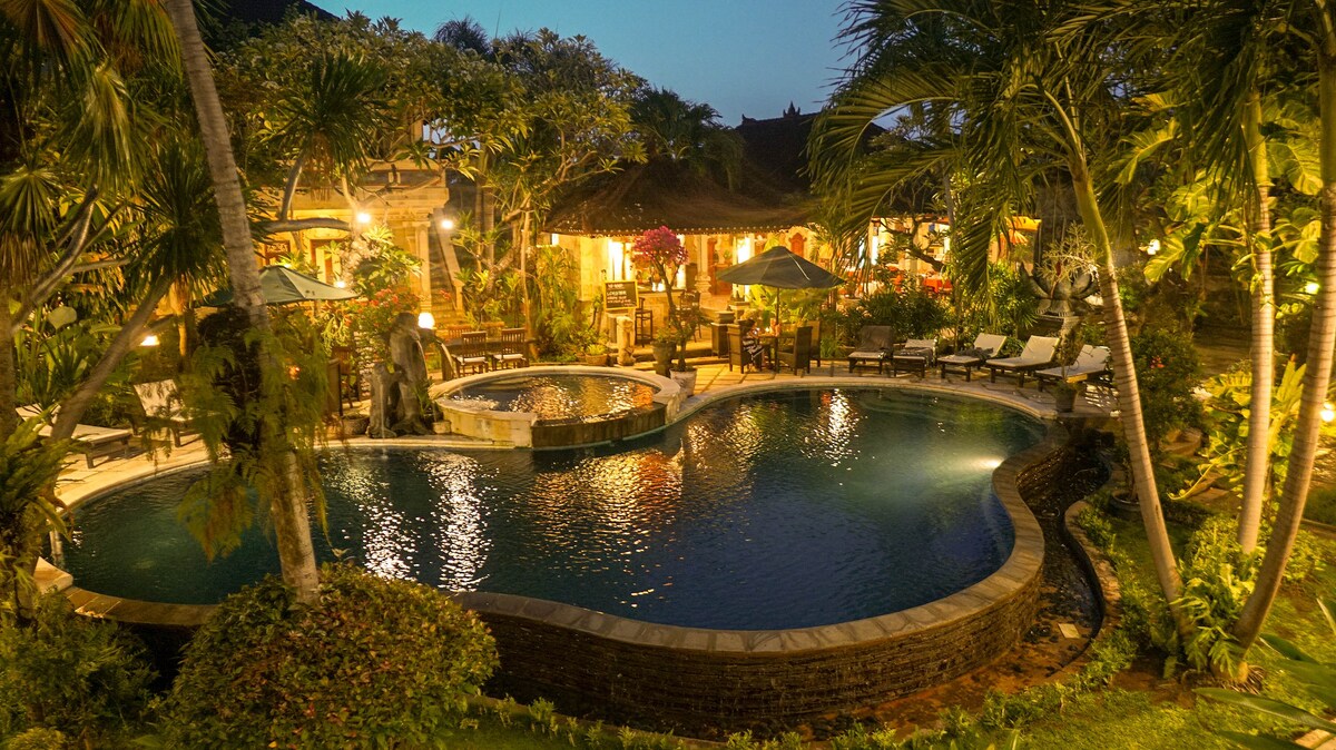 The outdoor area features a winding pool surrounded by lush greenery and illuminated by warm lighting. Lounge chairs are positioned nearby, creating spaces for relaxation. A tranquil atmosphere is enhanced by the surrounding tropical plants and the inviting design of the adjacent villa.