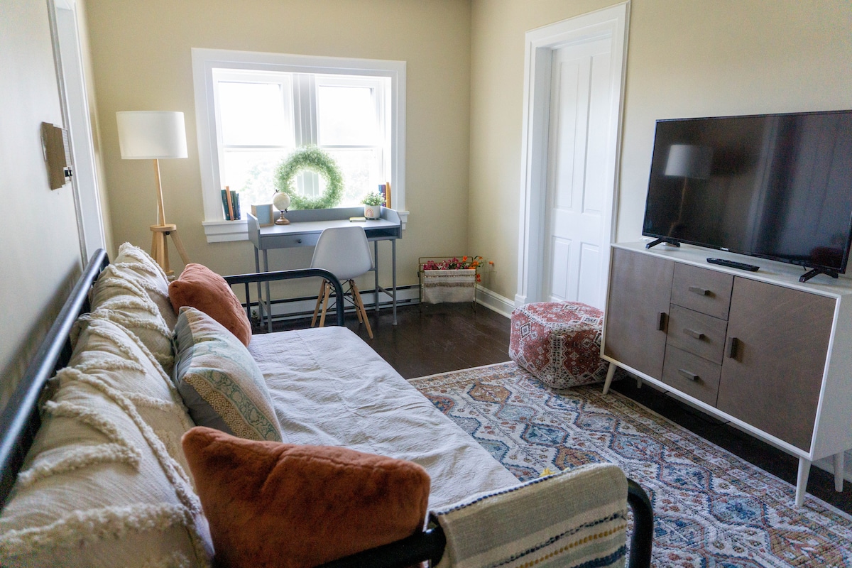 A cozy living area features a comfortable daybed adorned with soft pillows. A desk with a chair is positioned near a window, complemented by a decorative wreath. A television is situated on a light-colored media console, with a patterned rug adding warmth to the space.