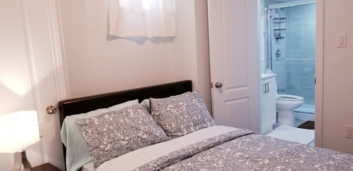 A cozy bedroom features a double size bed adorned with soft bedding and decorative pillows. Natural light enters through a window, illuminating the space. A doorway leads to an ensuite bathroom with modern fixtures, ensuring convenience for guests.